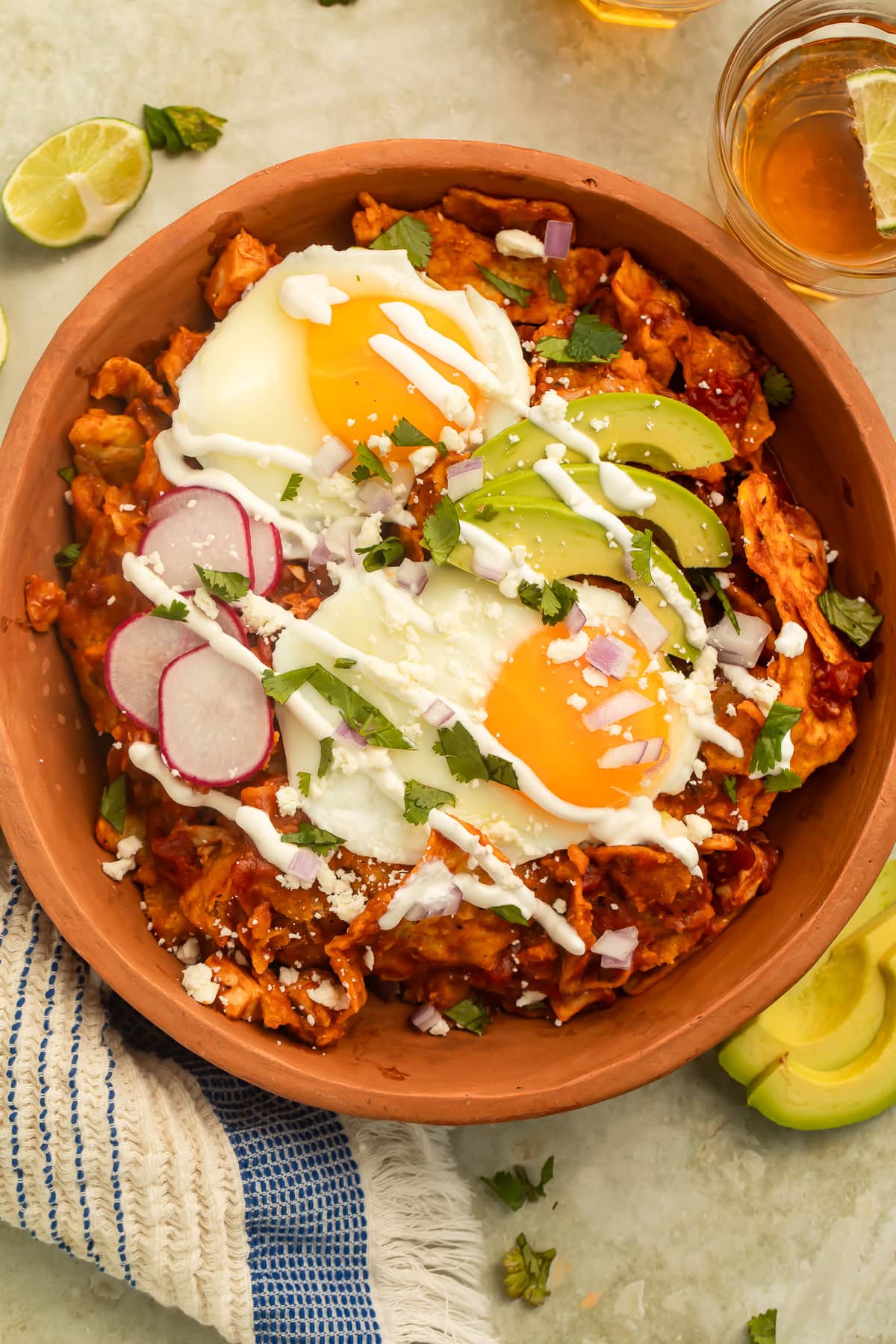 Bowl of red Chilaquiles topped with fried eggs, sliced avocado, sliced radish and a drizzle of crema.