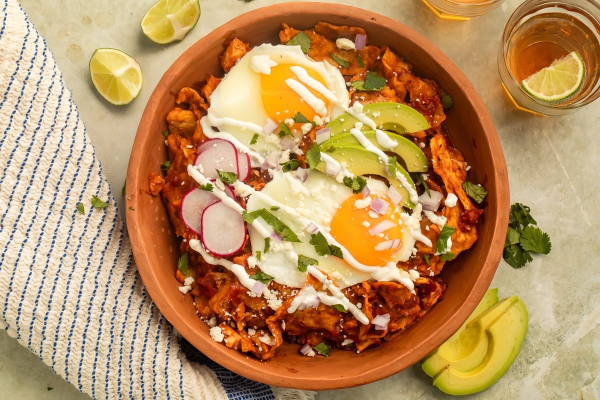 Terracotta bowl of red Chilaquiles topped with fried eggs, sliced avocado and a drizzle of crema. There are lime wedges and a glass of beer to one side.