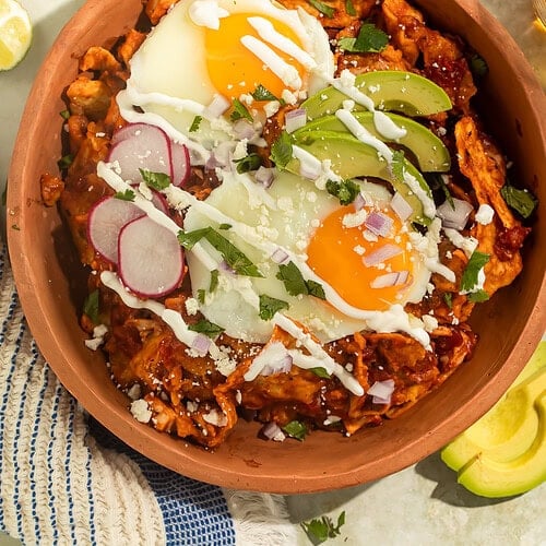 A terracotta bowl of Chilaquiles topped with fried eggs, sliced avocado, radishes and crema.
