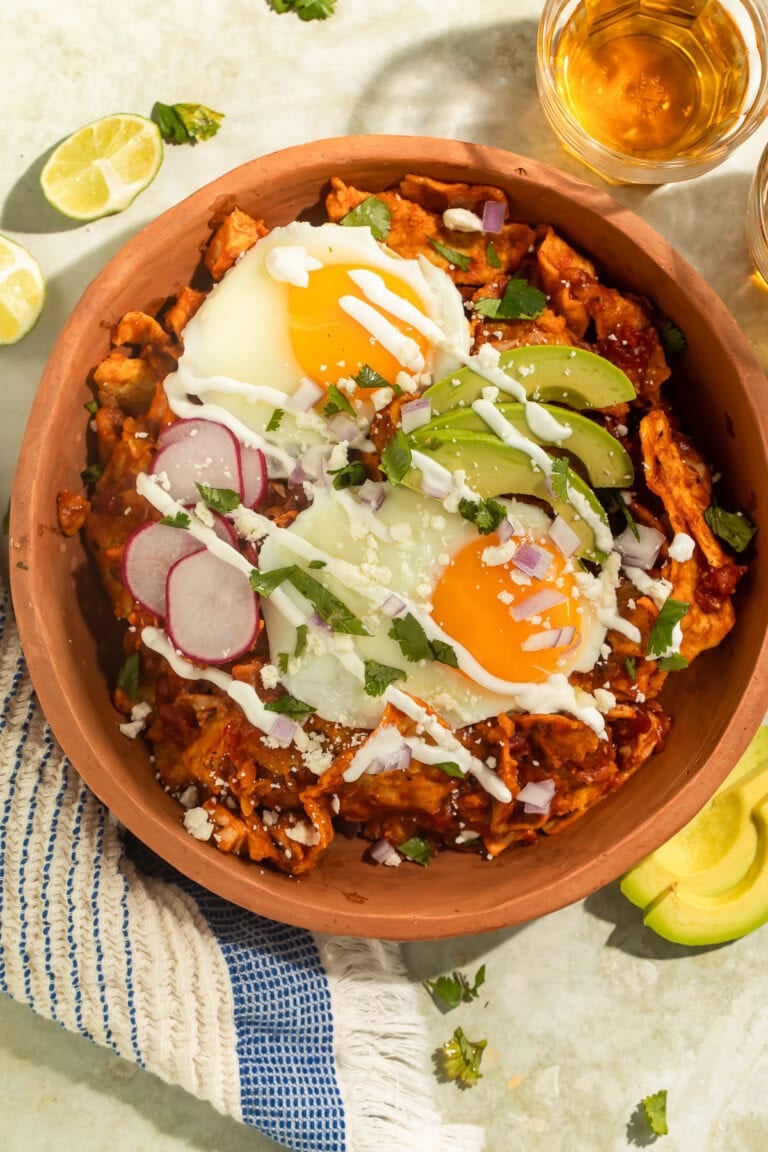 A terracotta bowl of Chilaquiles topped with fried eggs, sliced avocado, radishes and crema.