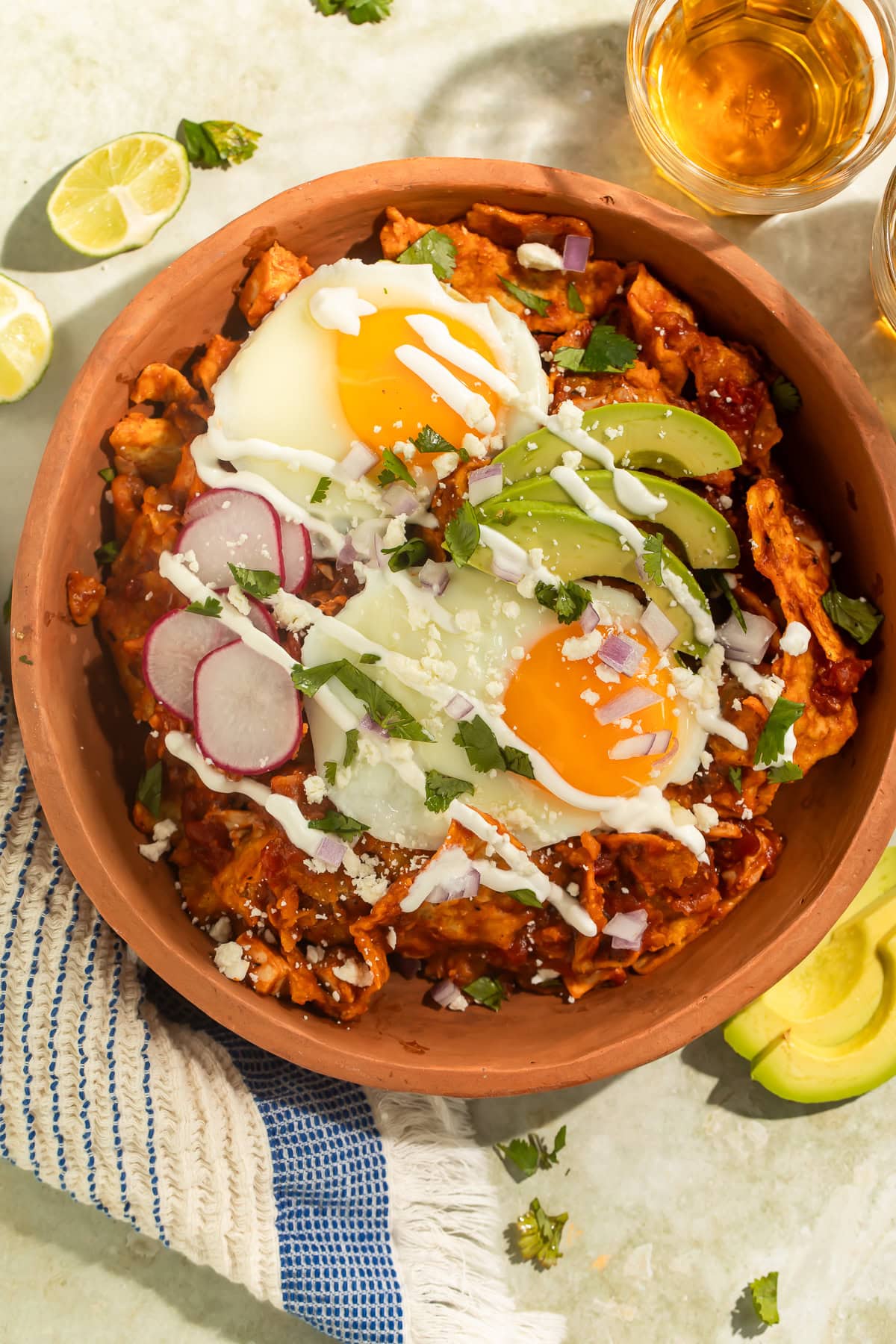 A terracotta bowl of Chilaquiles topped with fried eggs, sliced avocado, radishes and crema.