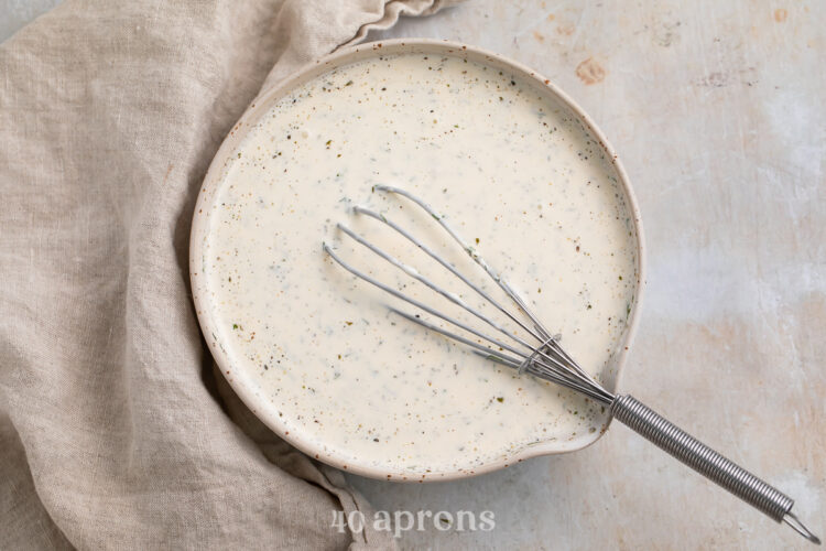 Overhead view of a bowl of buttermilk ranch dressing in a medium mixing bowl with a whisk on a neutral cloth napkin.