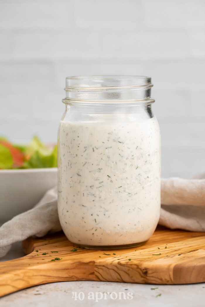 Side view of a mason jar filled with homemade buttermilk ranch dressing, sitting on a wooden serving board.