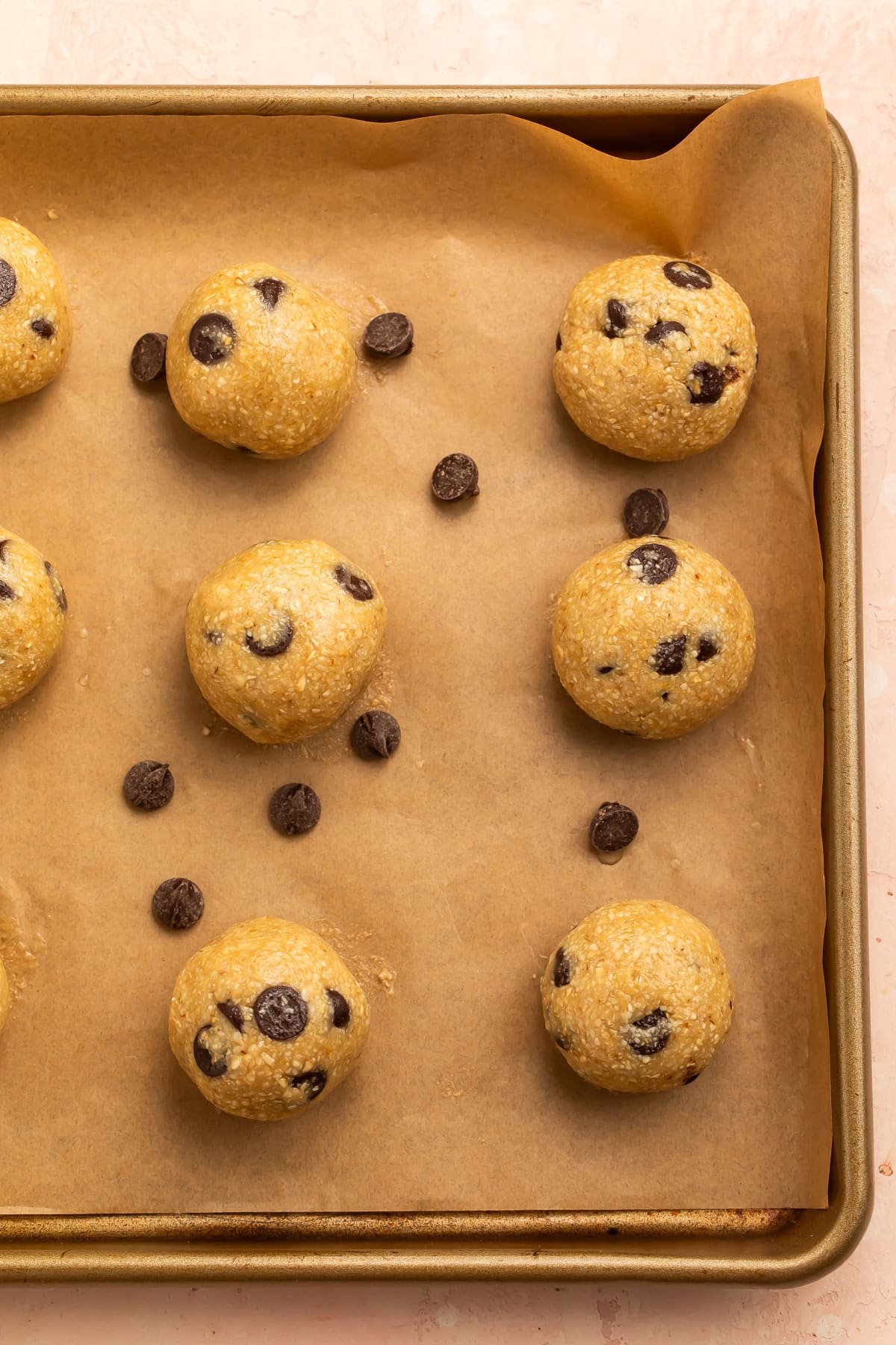 Raw chocolate chip cookie dough balls on a parchment lined baking sheet scattered with chocolate chips.