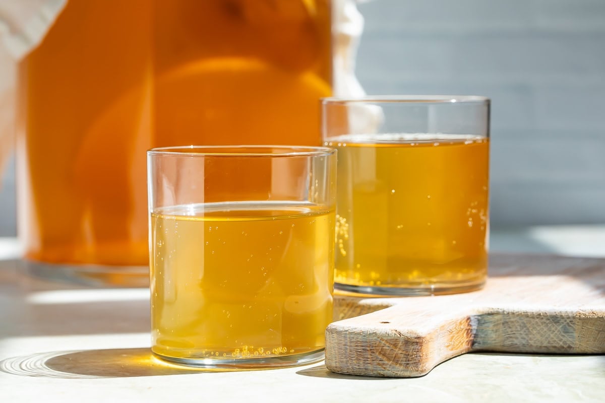 Close up of two glasses of kombucha with a jar of kombucha with a SCOBY in the background.