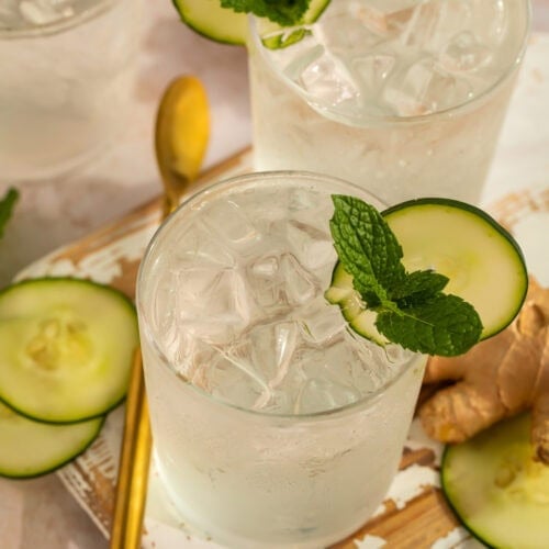 Two cucumber ginger mocktails garnished with mint leaves and cucumber discs on a wooden tray.