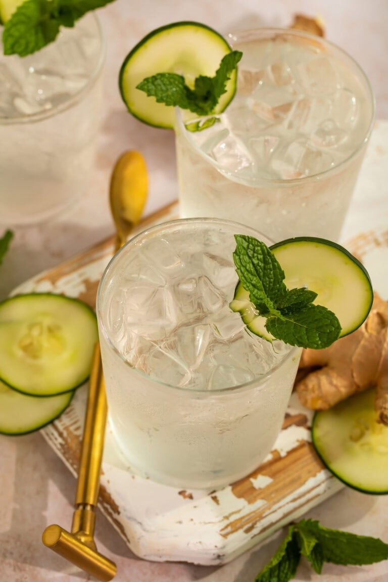Two cucumber ginger mocktails garnished with mint leaves and cucumber discs on a wooden tray.