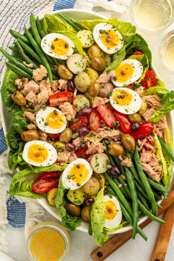 A large oval platter of Nicoise salad with potatoes, green beans, eggs, lettuce, olives, capers, tomatoes, and tuna.