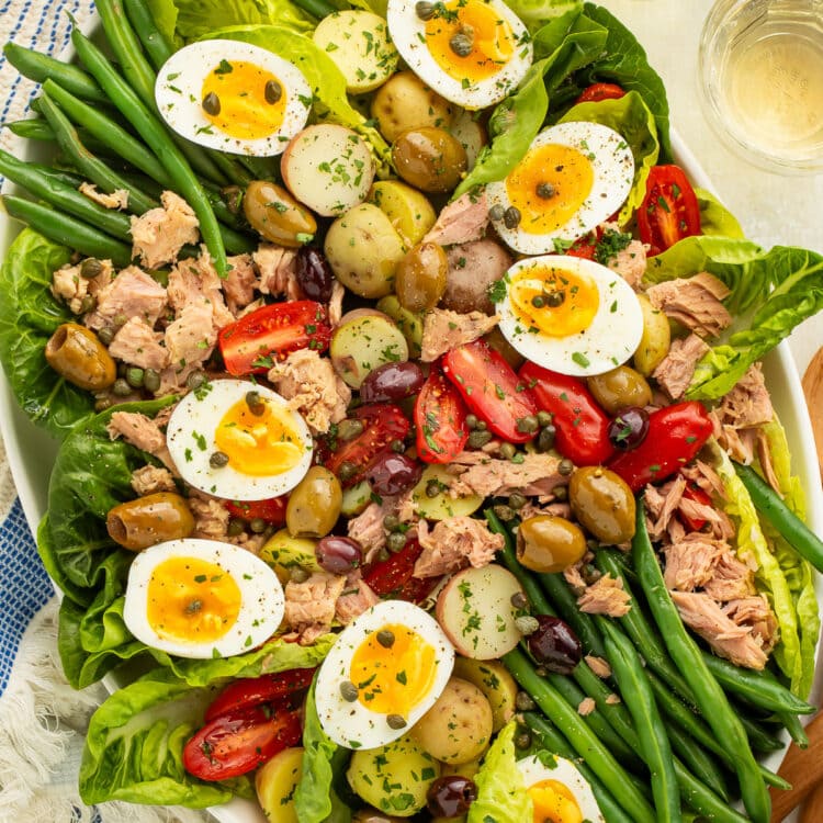 A large oval platter of Nicoise salad with potatoes, green beans, eggs, lettuce, olives, capers, tomatoes, and tuna.