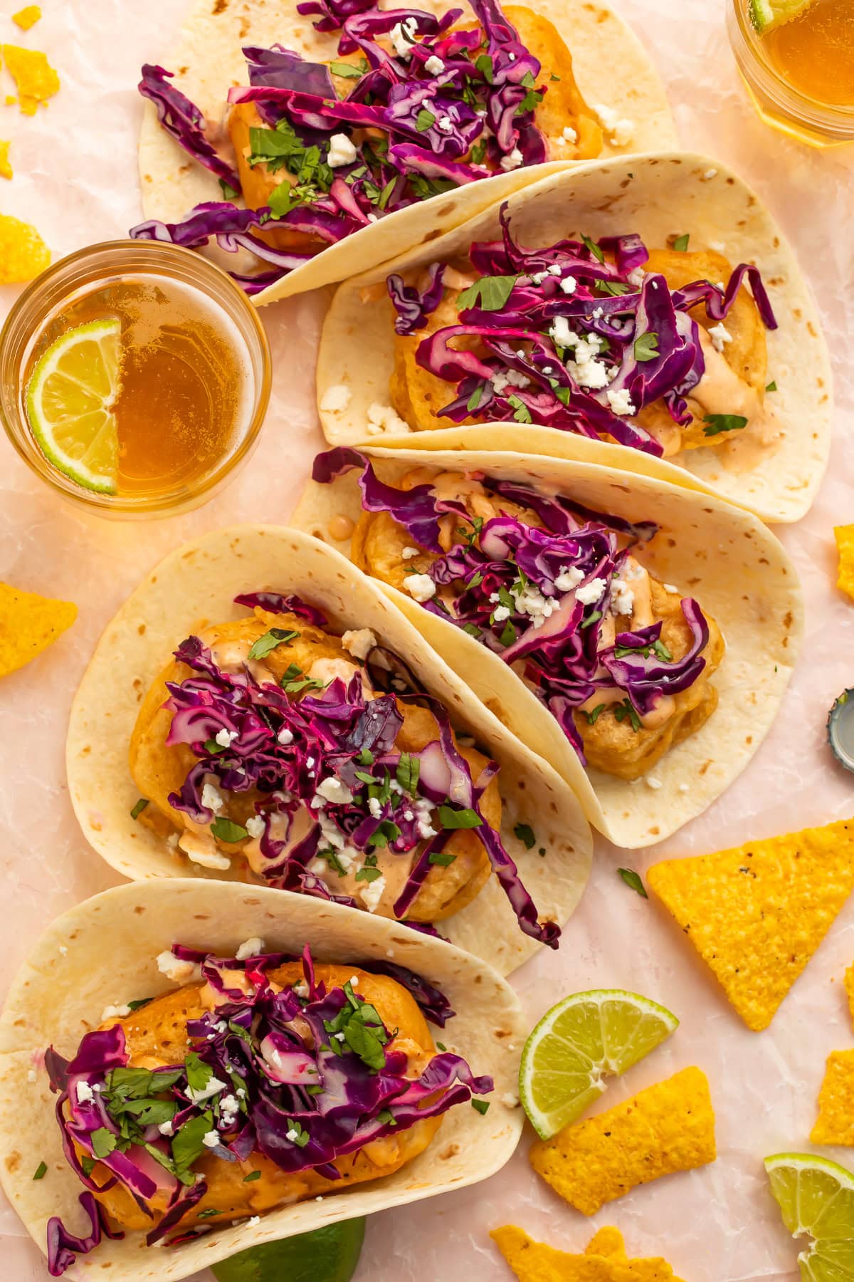 Four beer battered tofu tacos on a pink surface with tortilla chips, lime wedges, and a glass of beer