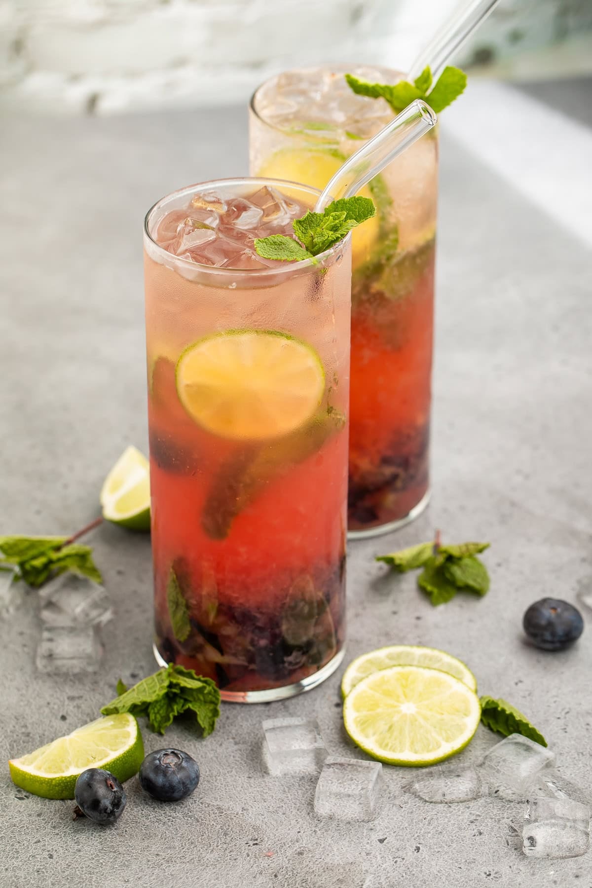 Side on view of two blueberry mojitos on a grey countertop surrounded by fresh mint, lime wheels and ice.