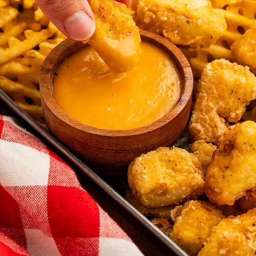 Hand dipping a tofu nugget in a creamy orange sauce.
