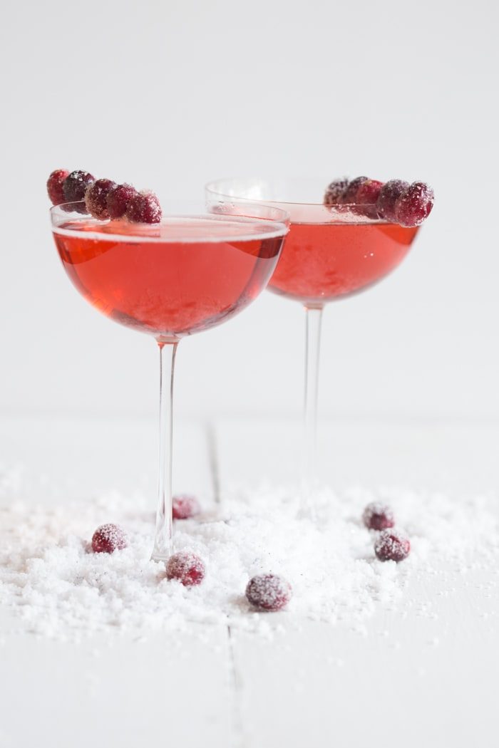Close-up of two cranberry champagne cocktails with sugar-coated cranberry garnishes and scattered sugared cranberries.