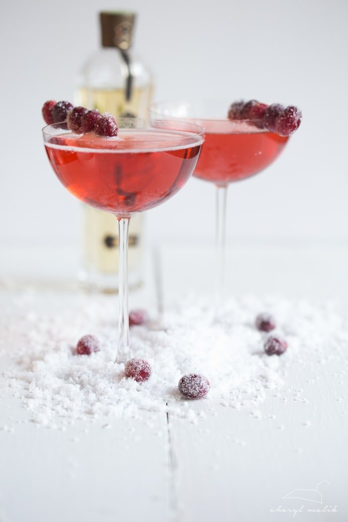 Two cranberry elderflower champagne cocktails in coupe glasses with sugared cranberry garnishes on snowy surface.