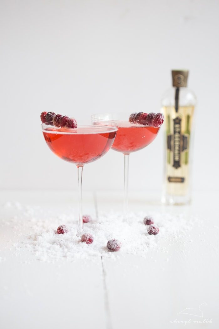 Two cranberry champagne cocktails with sugared cranberry garnishes and St. Germain elderflower liqueur bottle.