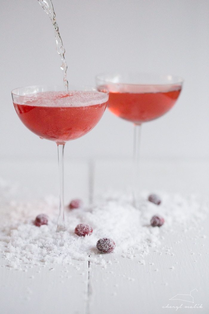 Champagne being poured into cranberry cocktail creating bubbles and fizz in coupe glass on snowy surface.