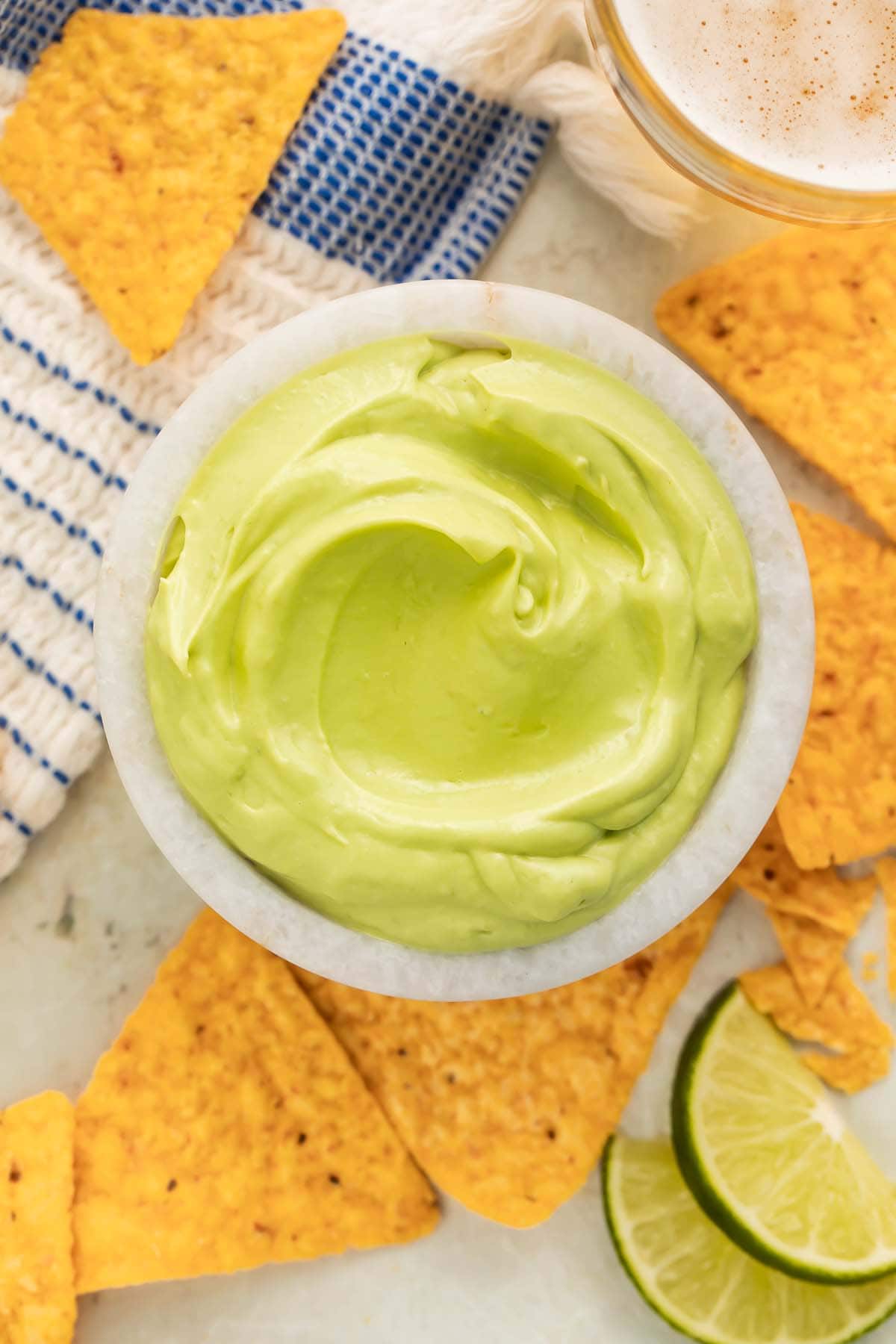 Overhead view of a small bowl of avocado crema on a table surrounded by triangle-shaped tortilla chips and a lime wedge.