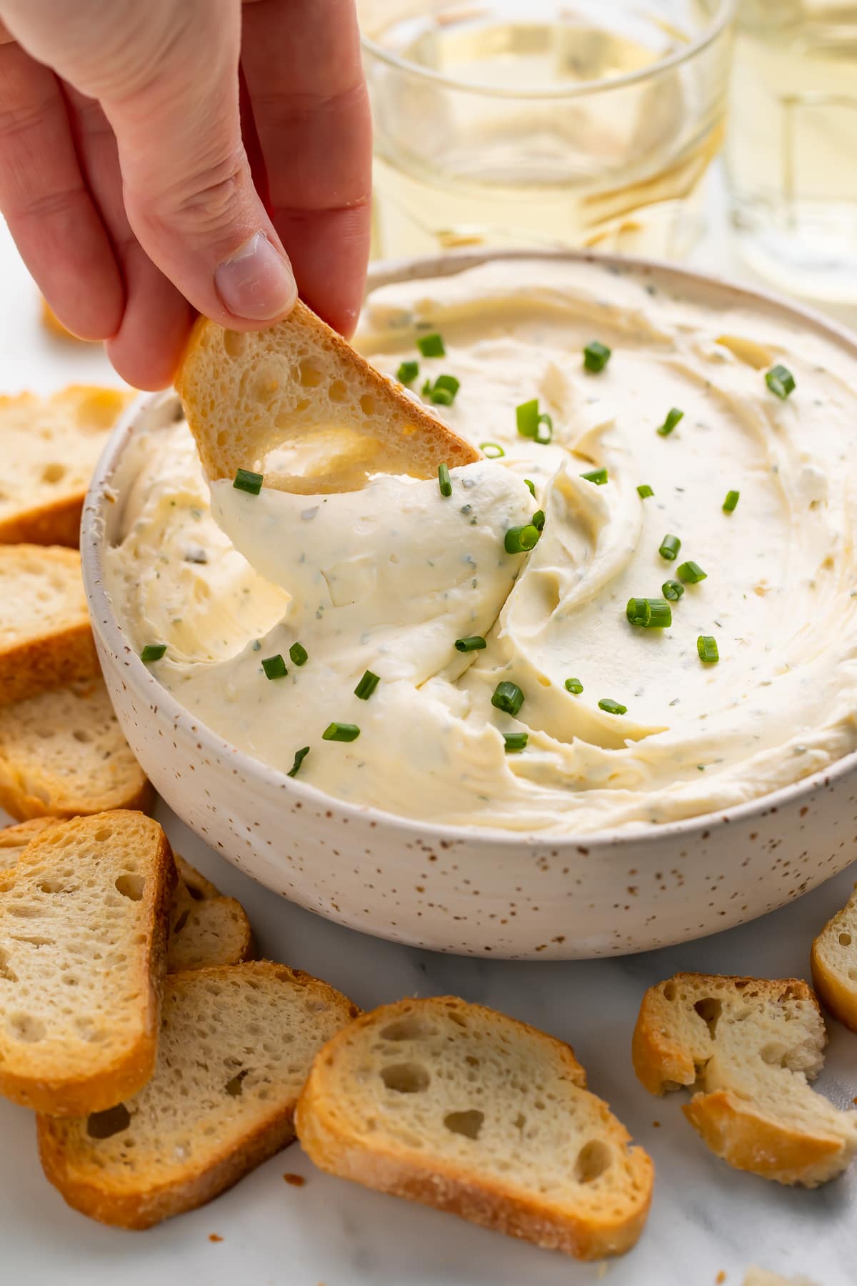 A white woman's hand dipping a piece of crostini into a bowl of creamy homemade boursin cheese.