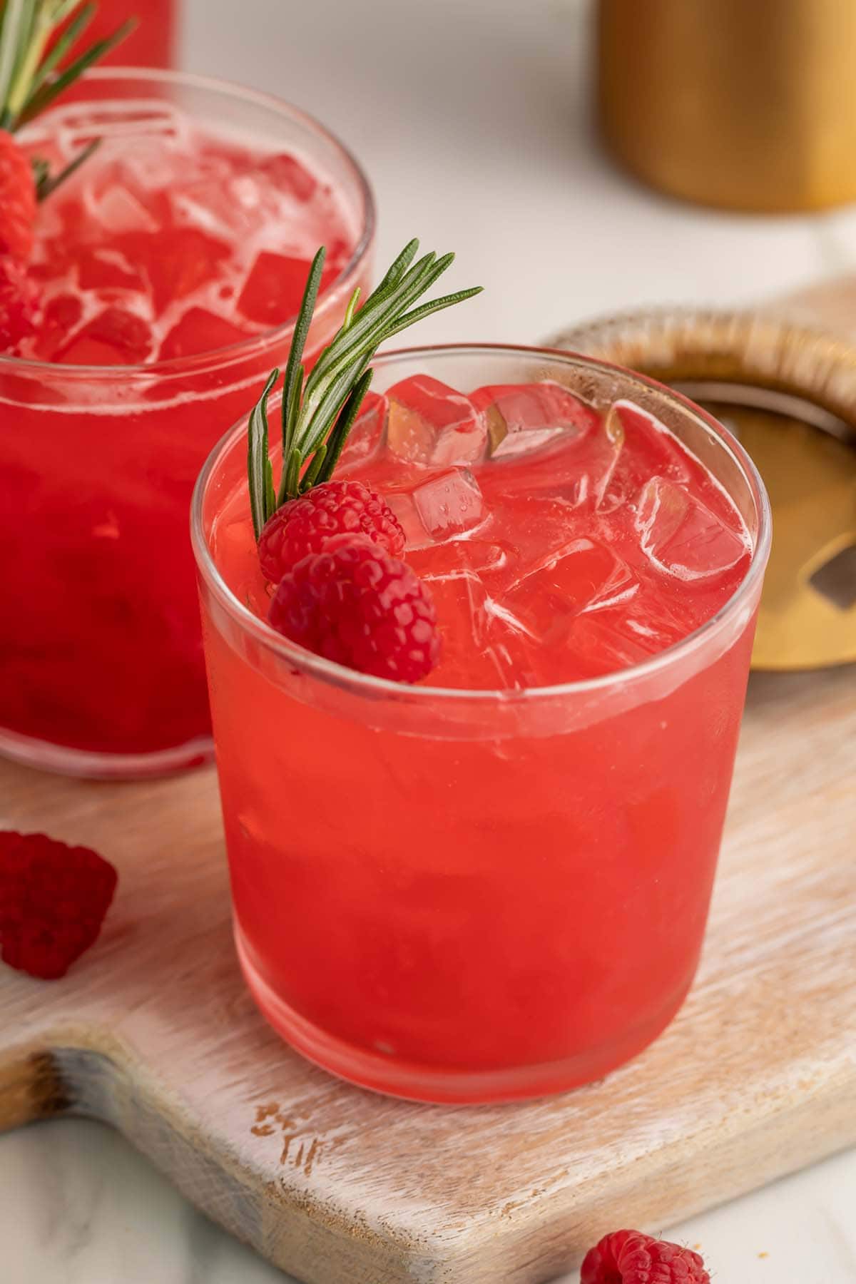 A bright red raspberry cocktail in a glass, garnished with a raspberry and sprig of rosemary.