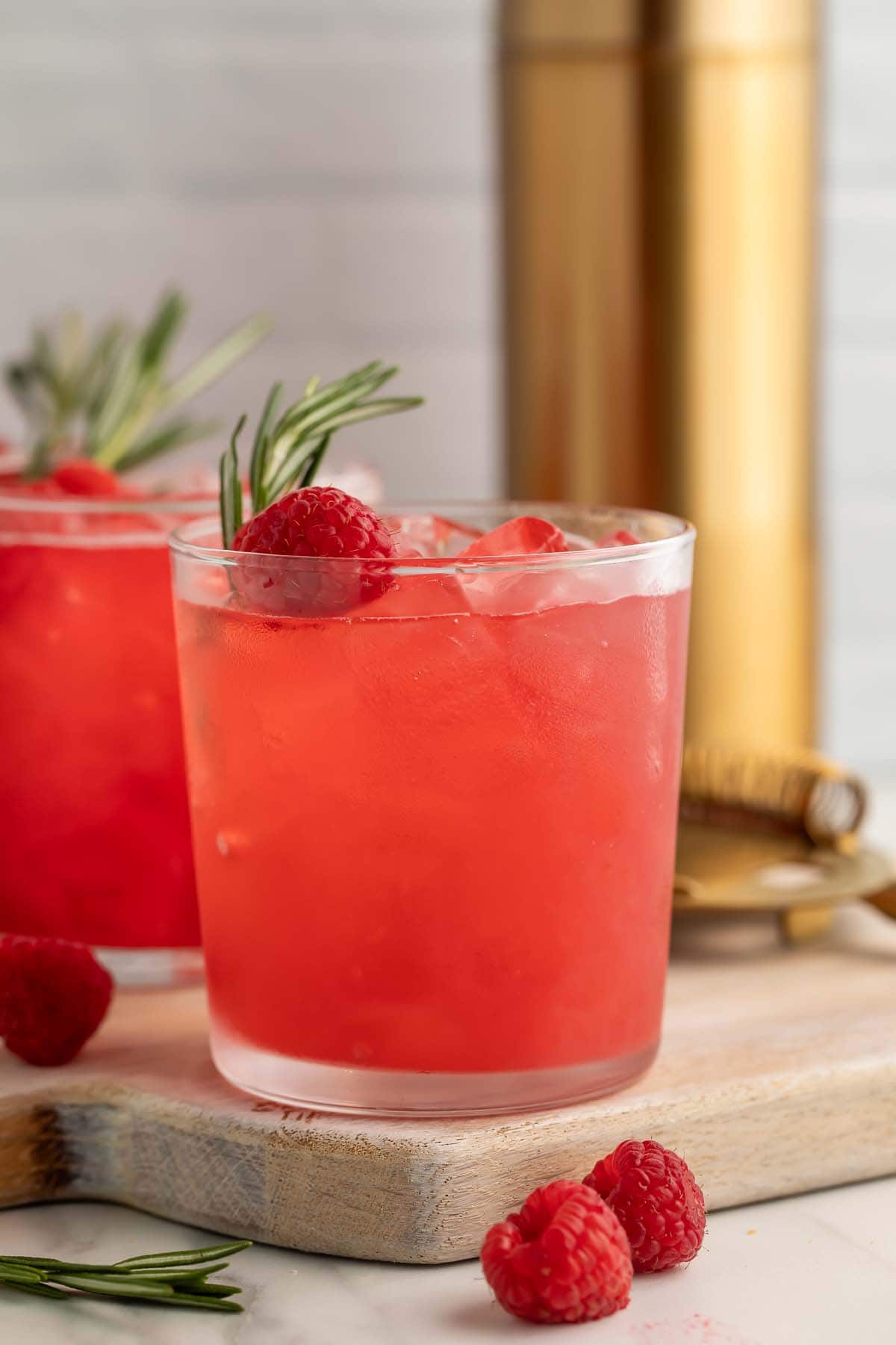 A bright red raspberry cocktail in a glass, garnished with a raspberry and sprig of rosemary.