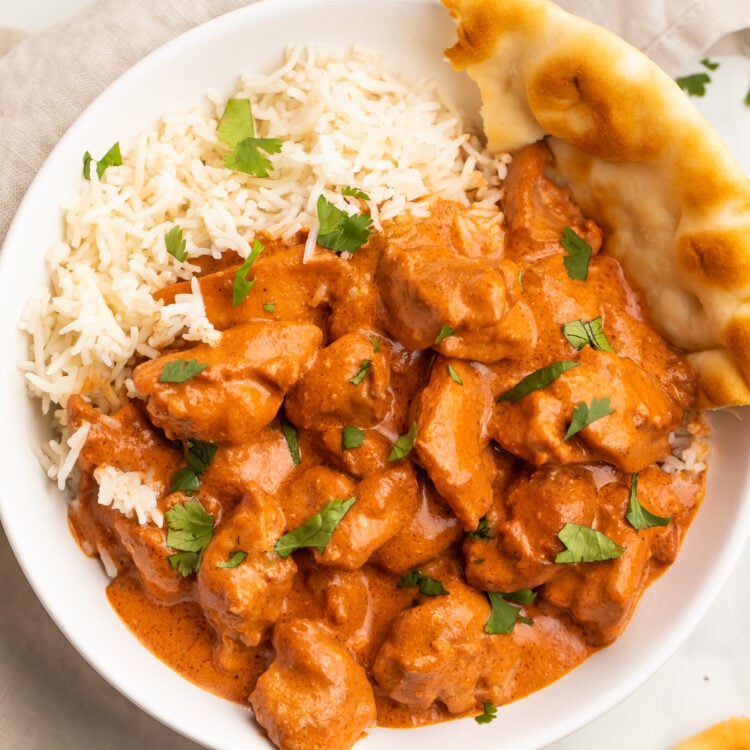 Zoomed out photo of butter chicken in a creamy red-orange sauce plated next to white rice and naan.