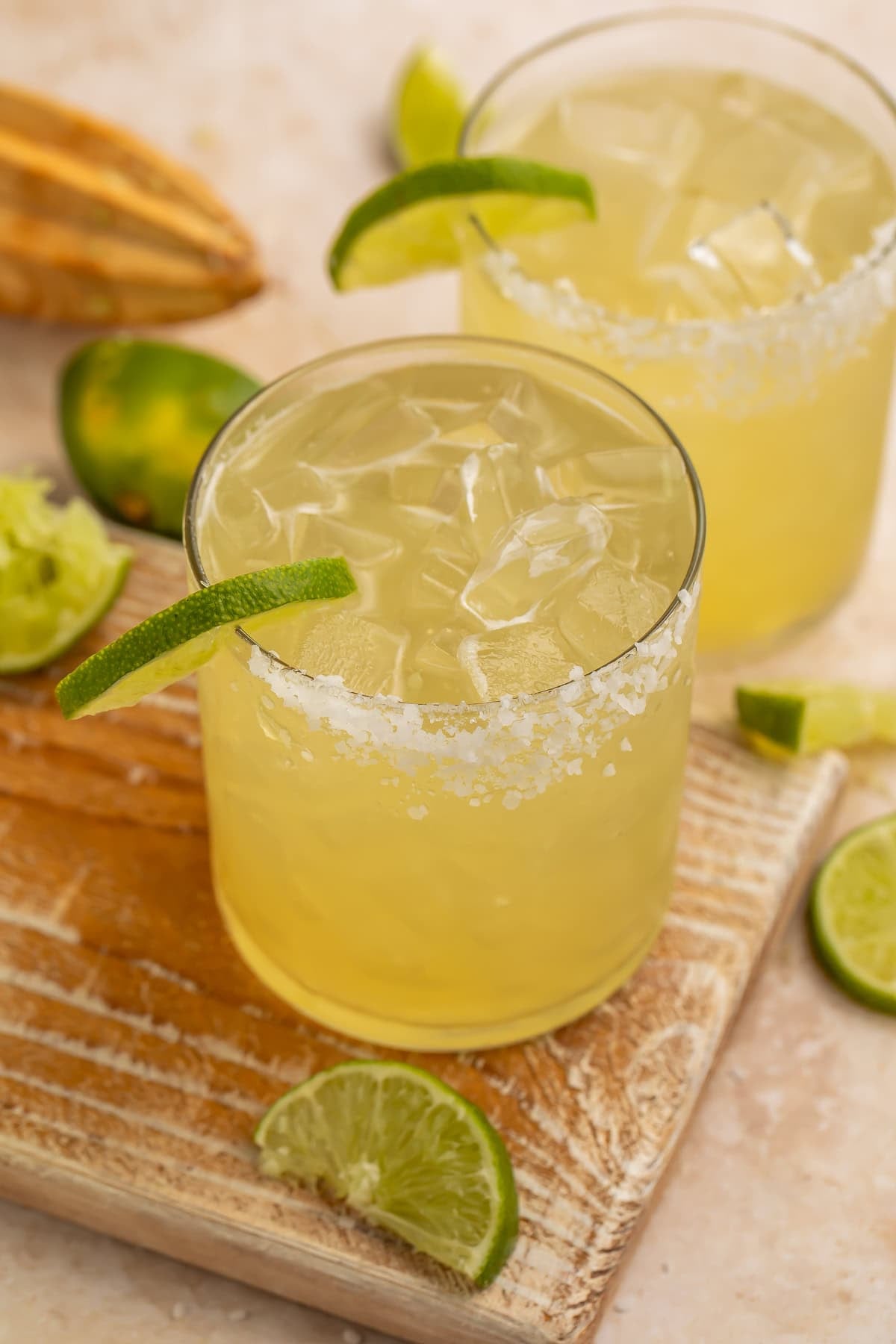 Close up of two margaritas with lime wedges and a salt rim. spent lime halves and a wooden reamer are just visible in the background.
