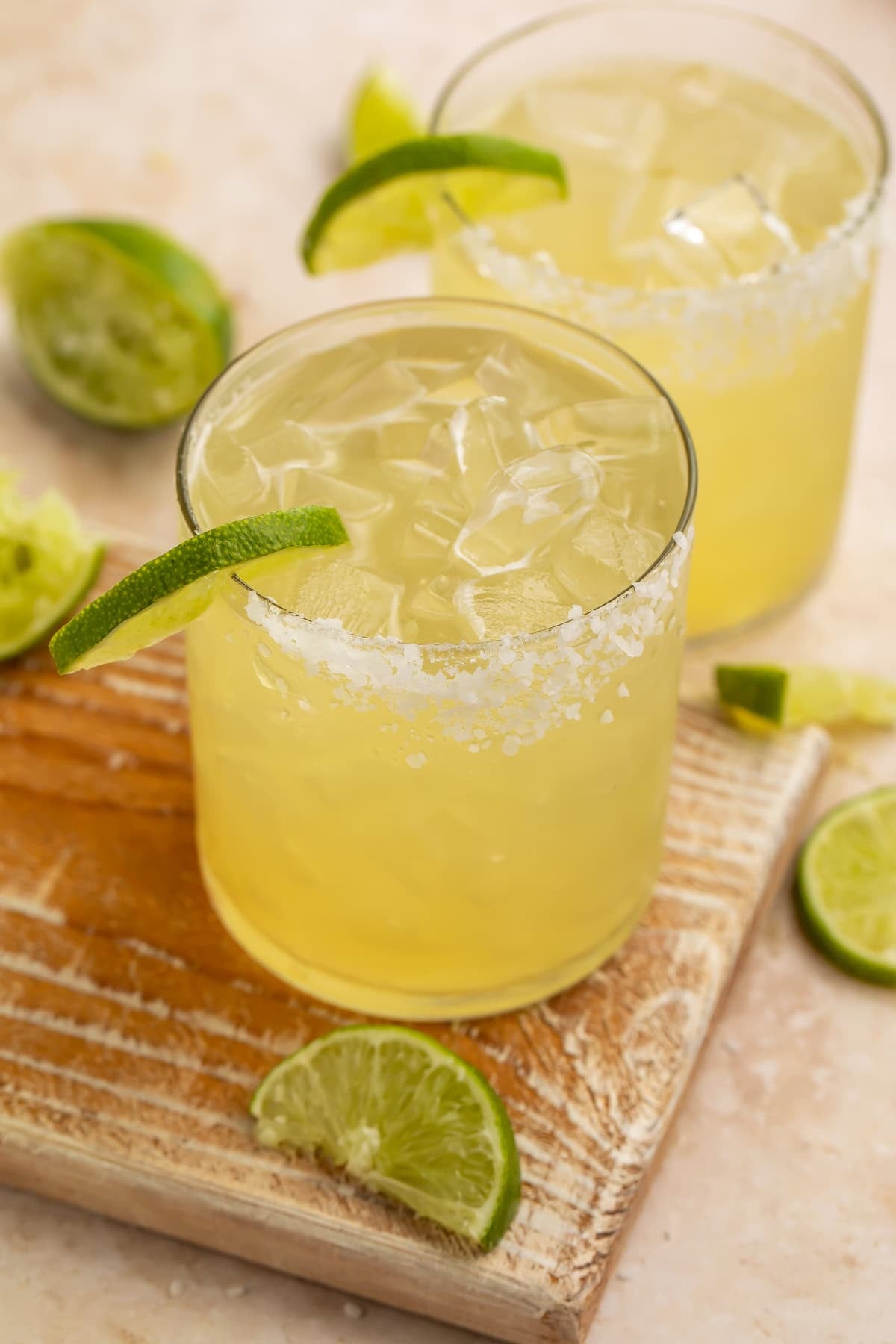 Two margaritas on a wooden board with plenty of ice, lime wedges and generous salt rims.