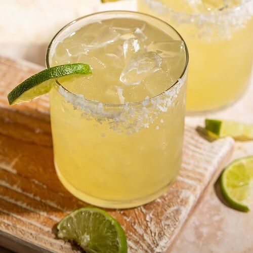 A pair of margaritas with a lime wedge and a salt rim on a wooden board.