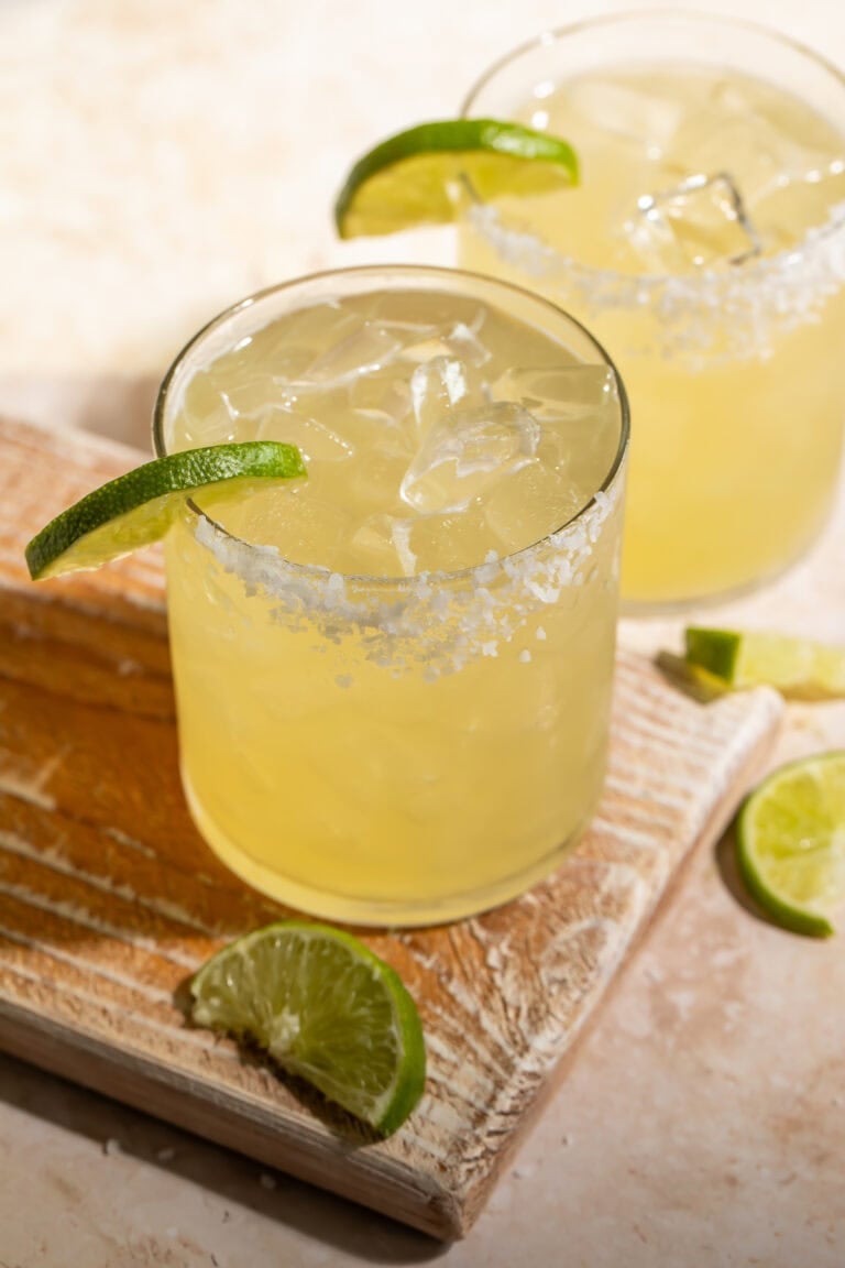 A pair of margaritas with a lime wedge and a salt rim on a wooden board.