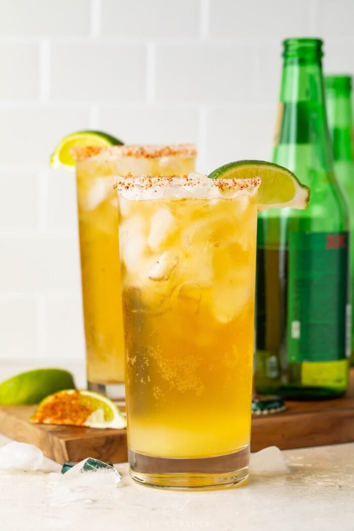 Two tall salt-rimmed glasses of chelada in front of green beer bottles