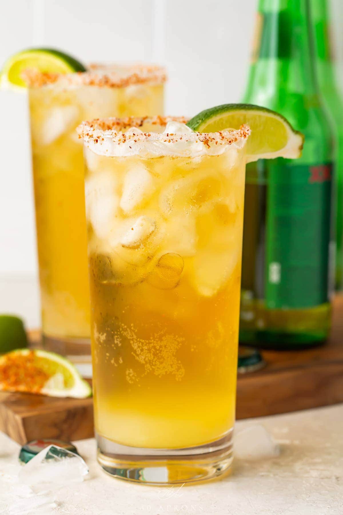 Close up of two tall salt-rimmed glasses of chelada in front of green beer bottles
