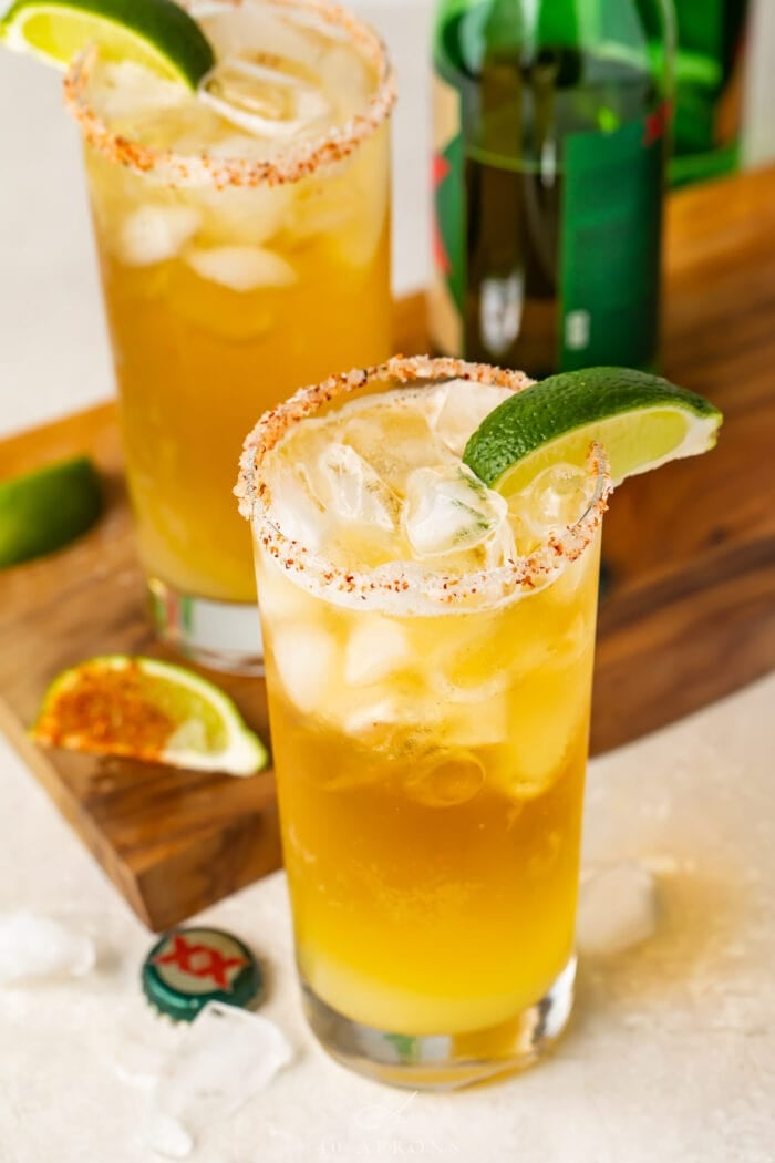 Tall salt-rimmed glass of chelada with lime wedge garnish on a table with a wooden board