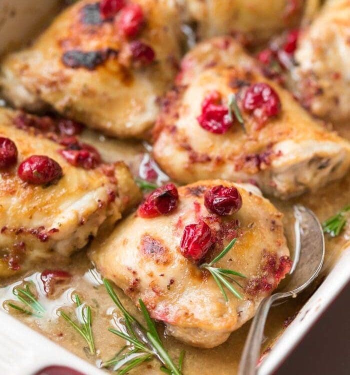 Paleo cranberry rosemary one pan chicken thighs in a baking dish with vintage spoon topped with cranberries and fresh rosemary