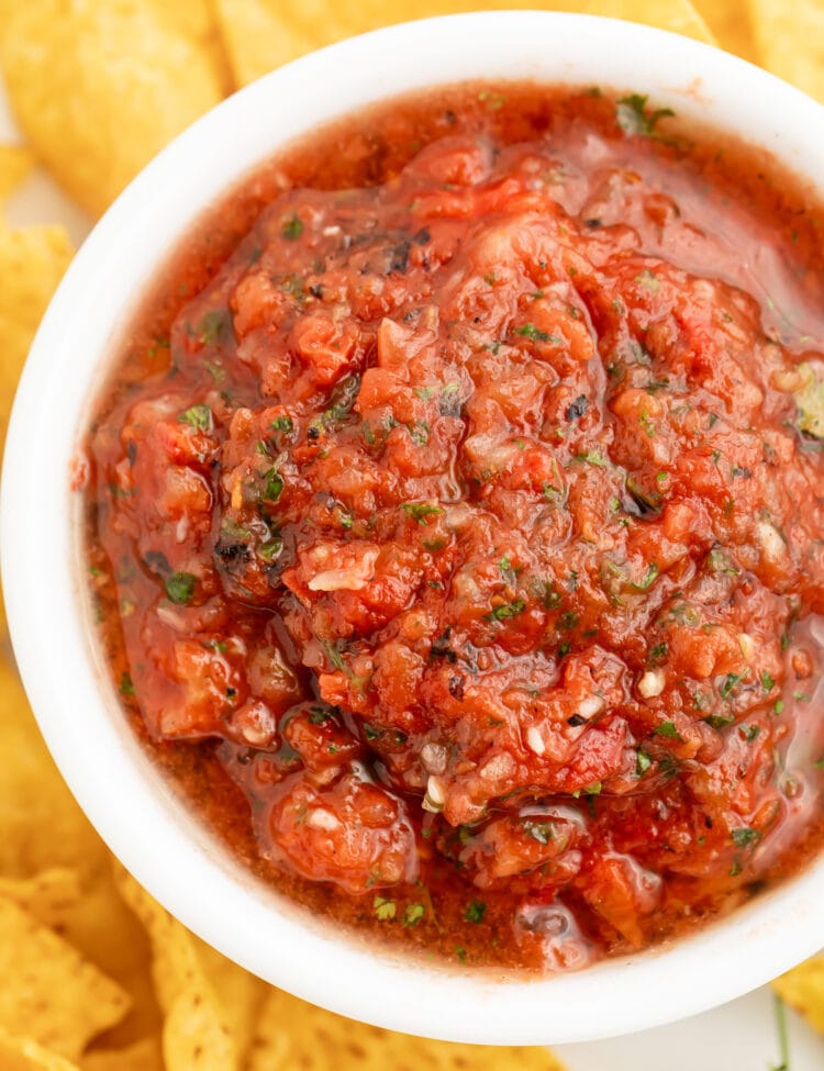 Top-down, overhead view of a bowl of deep red, slightly chunky restaurant-style salsa in a white bowl surrounded by tortilla chips.