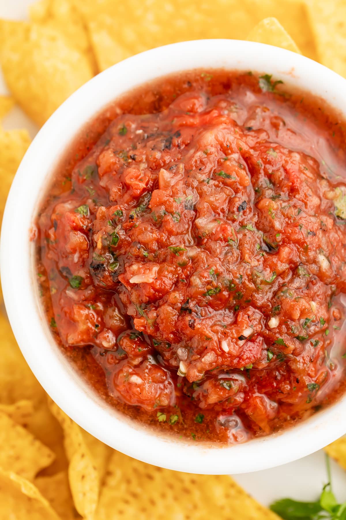 Top-down, overhead view of a bowl of deep red, slightly chunky restaurant-style salsa in a white bowl surrounded by tortilla chips.