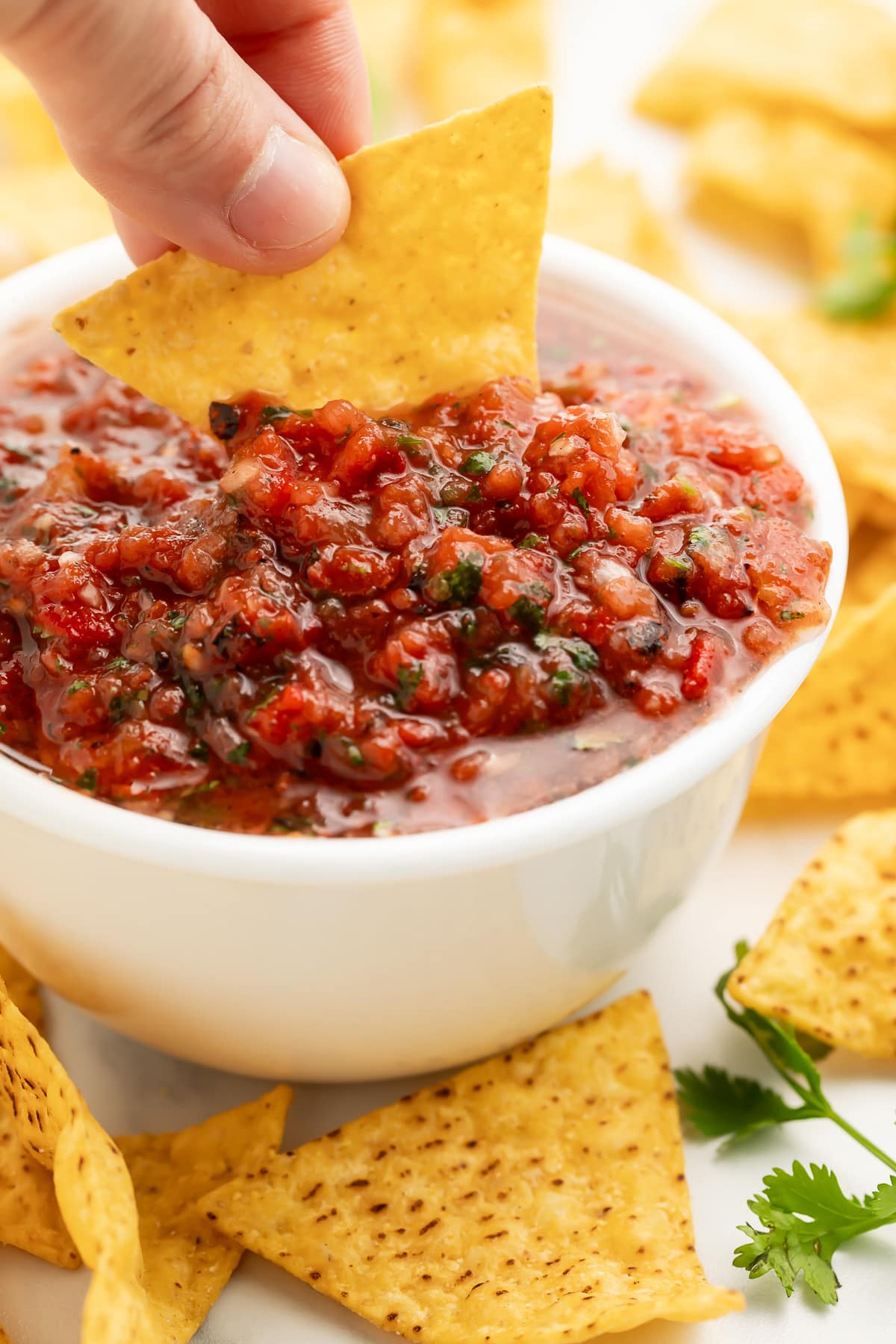 A white woman's hand holding a tortilla chip and using it to scoop red, chunky salsa out of a white bowl.