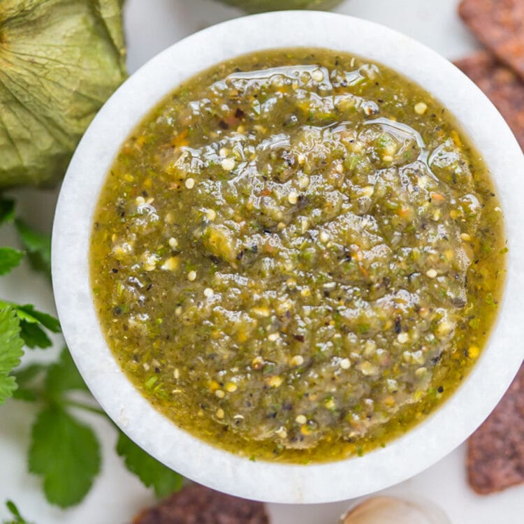 Top-down, overhead view of a small bowl of green roasted salsa verde surrounded by blue corn chips.