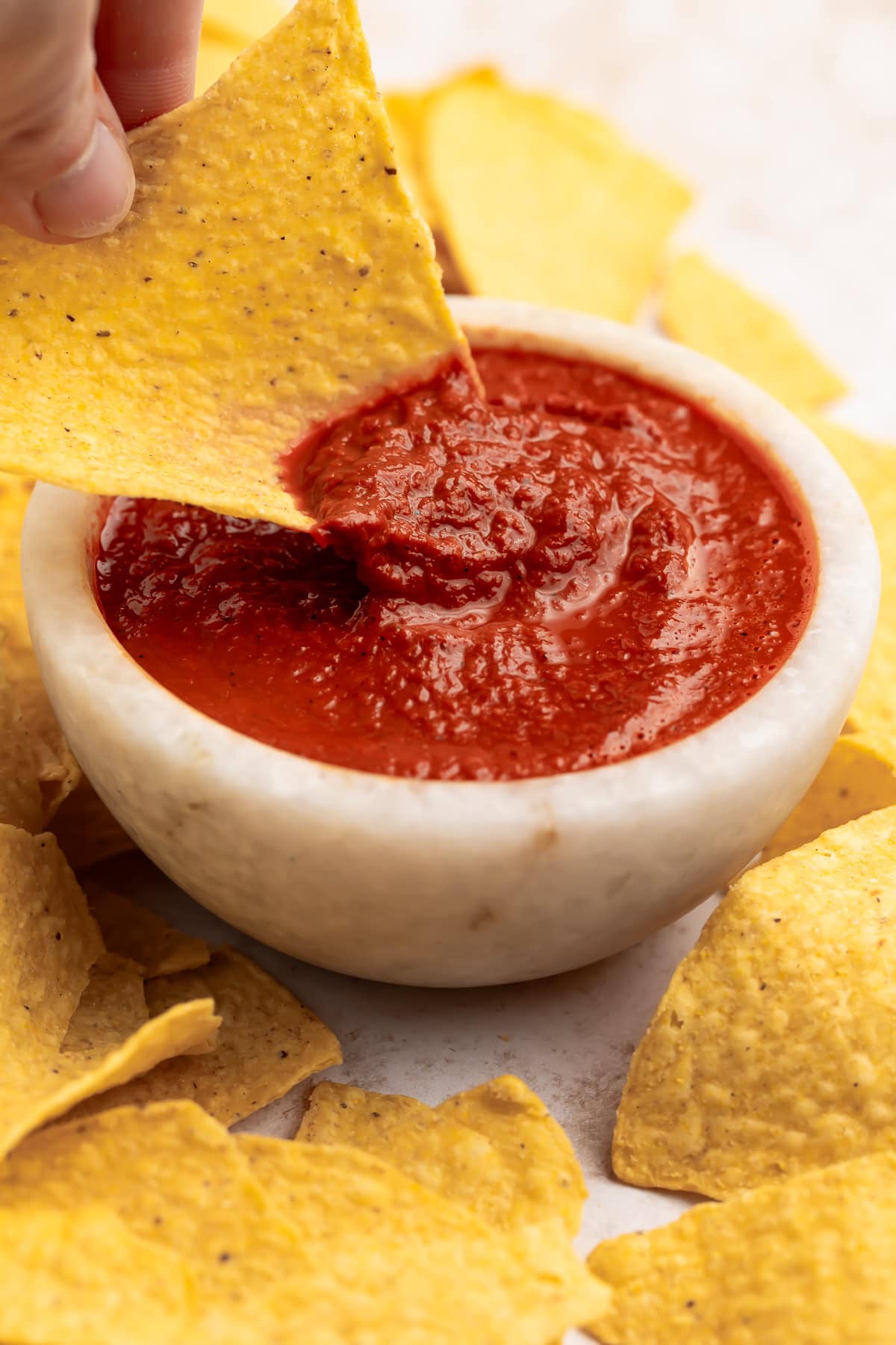 A white woman's hand holding a yellow corn tortilla as she uses the chip to scoop red guajillo salsa out of a bowl.