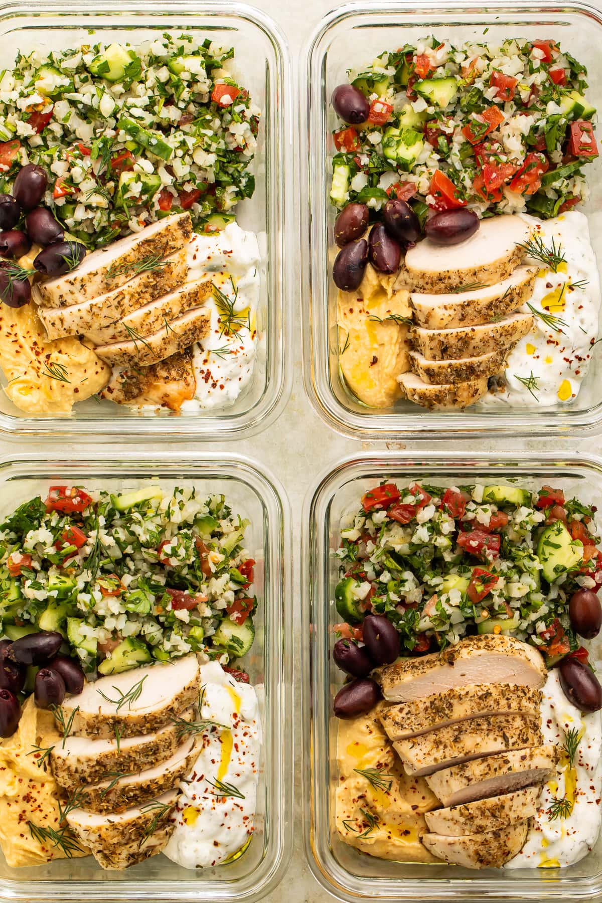 Close up of four glass meat prep containers of cauliflower tabbouleh salad, sliced chicken breast, olives and yogurt sauce.