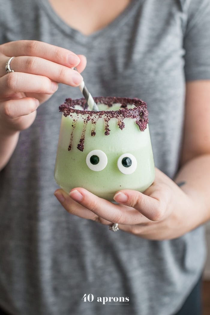 Someone holding Monsteritas green Halloween cocktails with purple dripping rim and candy monster eyes in glasses with a black and white straw