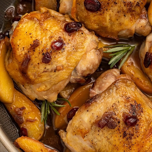 Golden-brown chicken thighs with dried cranberries and apple slices in a dark skillet with fresh rosemary.