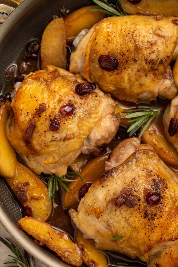 Golden-brown chicken thighs with dried cranberries and apple slices in a dark skillet with fresh rosemary.