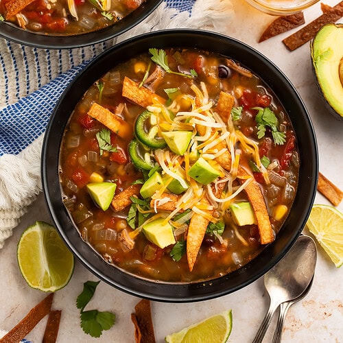 Two bowls of turkey tortilla soup with toppings on table with beer, lime wedges, and scattered tortilla strips