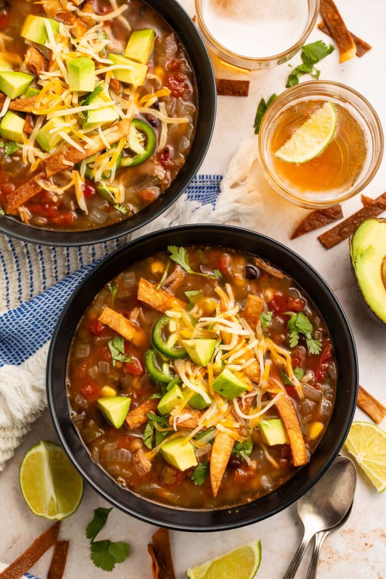 Two bowls of turkey tortilla soup with toppings on table with beer, lime wedges, and scattered tortilla strips