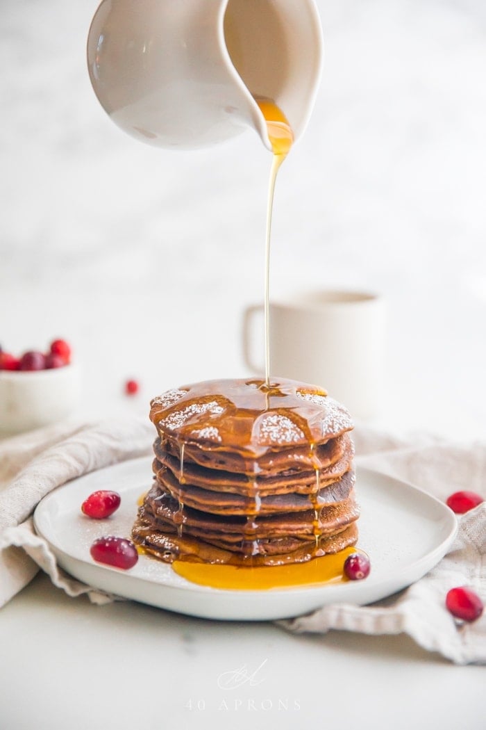 Stack of paleo gingerbread pancakes on a white plate with maple syrup pouring down the sides and fresh cranberries as garnish