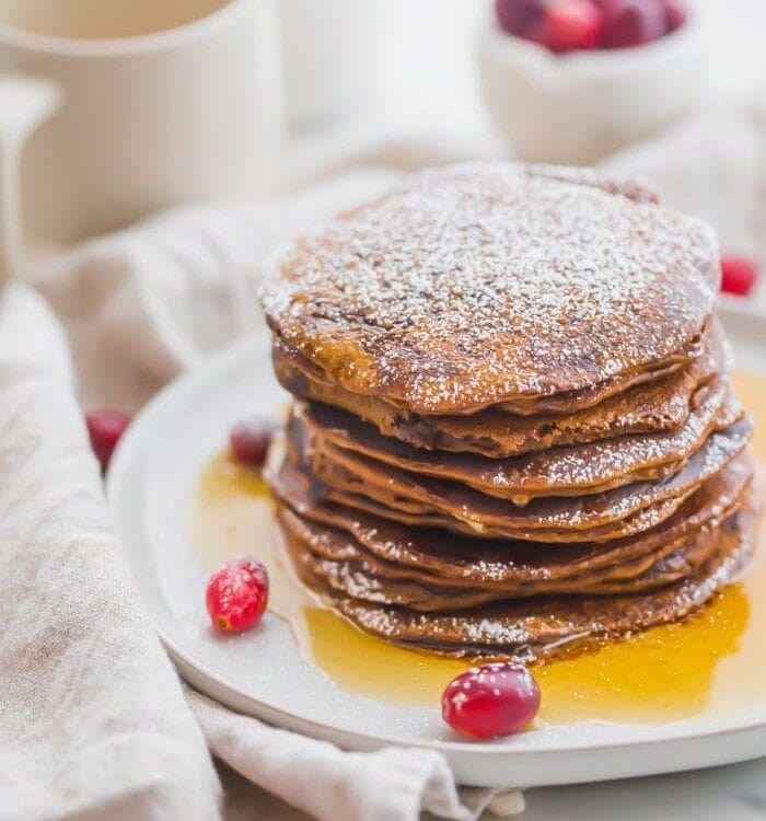 Stack of paleo gingerbread pancakes on a white plate with maple syrup around the bottom and fresh cranberries as garnish