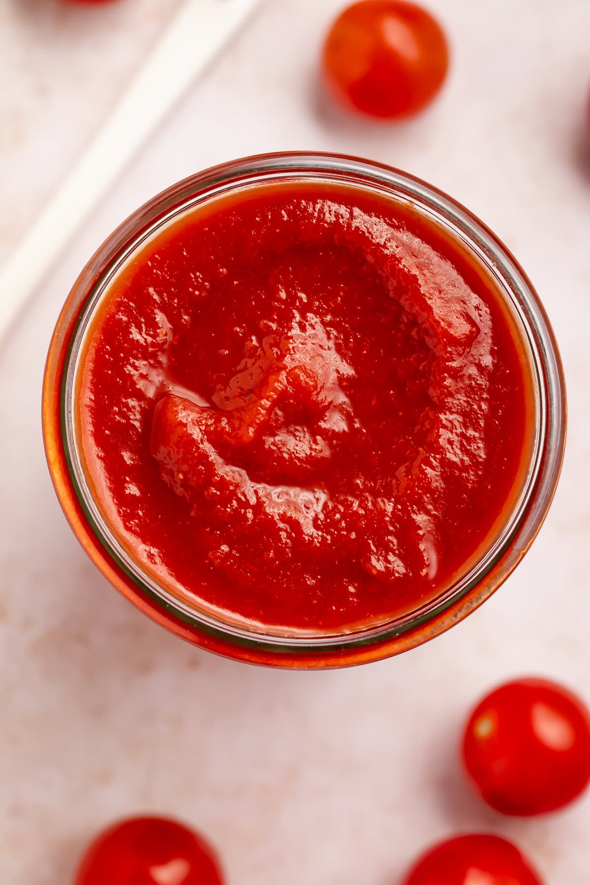Overhead photo of homemade ketchup in a glass jar with grape tomatoes scattered around it on the countertop. 