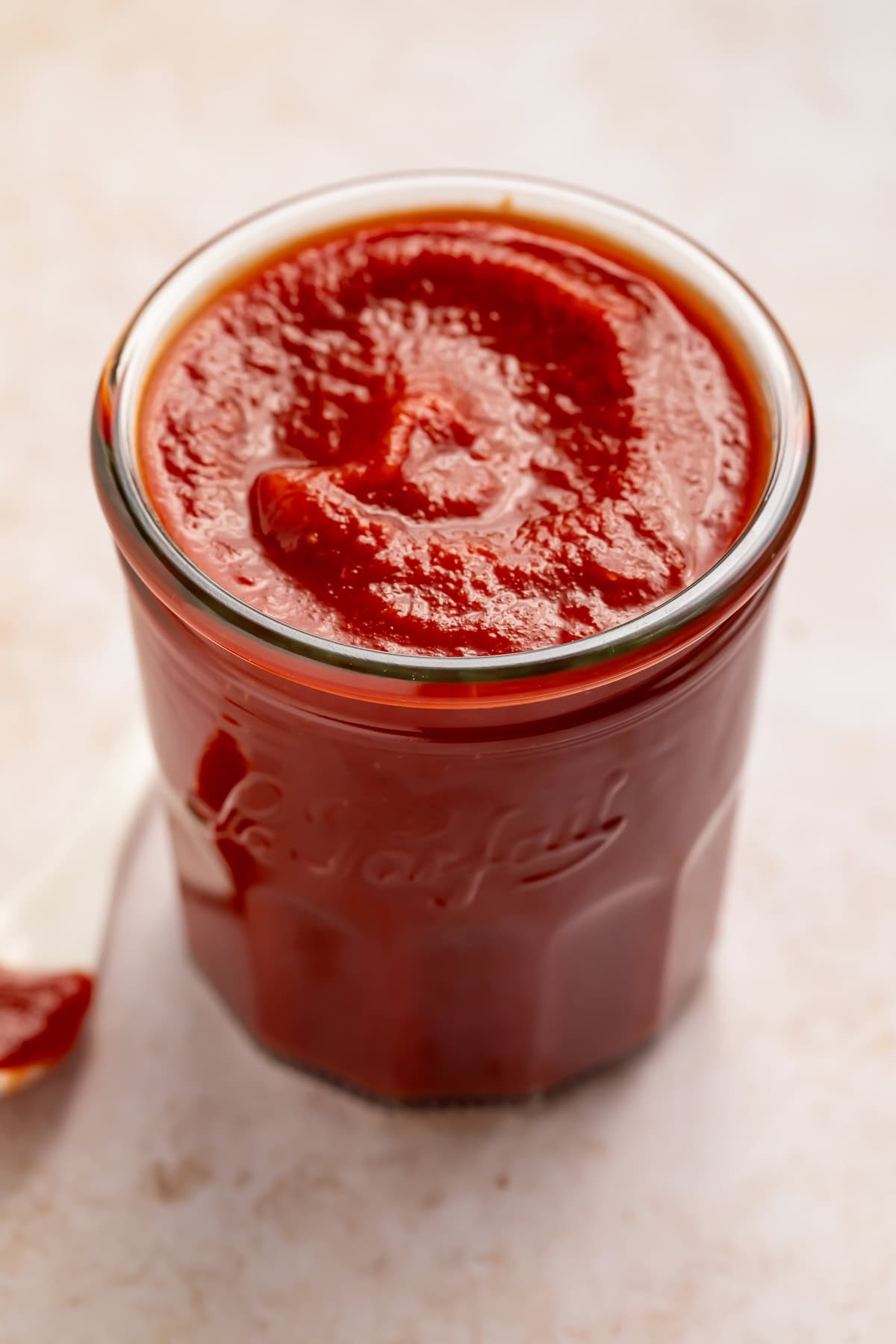 Glass jar of homemade ketchup with a white ketchup-dipped spoon off to one side. 