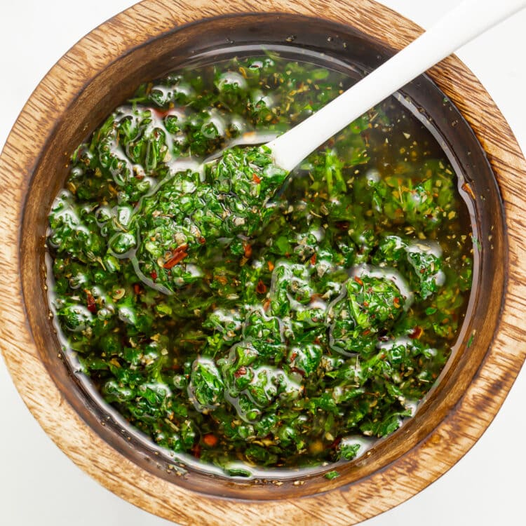 Zoomed out overhead photo of vibrant green chimichurri sauce in a small wooden bowl with a white spoon.