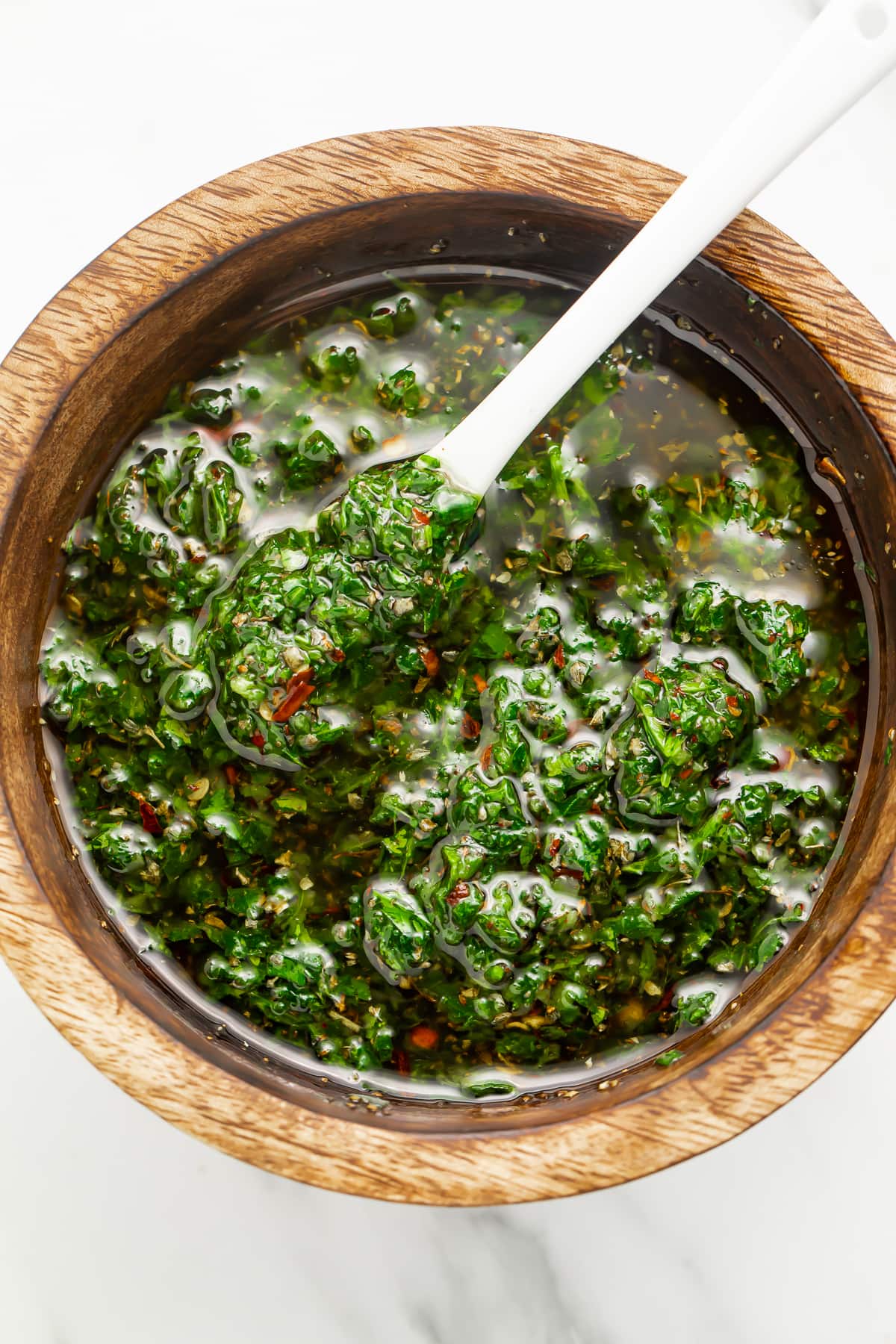 Zoomed out overhead photo of vibrant green chimichurri sauce in a small wooden bowl with a white spoon.